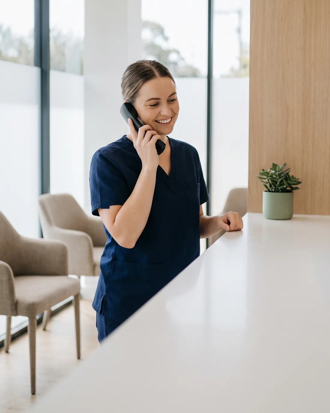 Dental receptionist in navy scrubs smiling as she answers the phone at the front desk